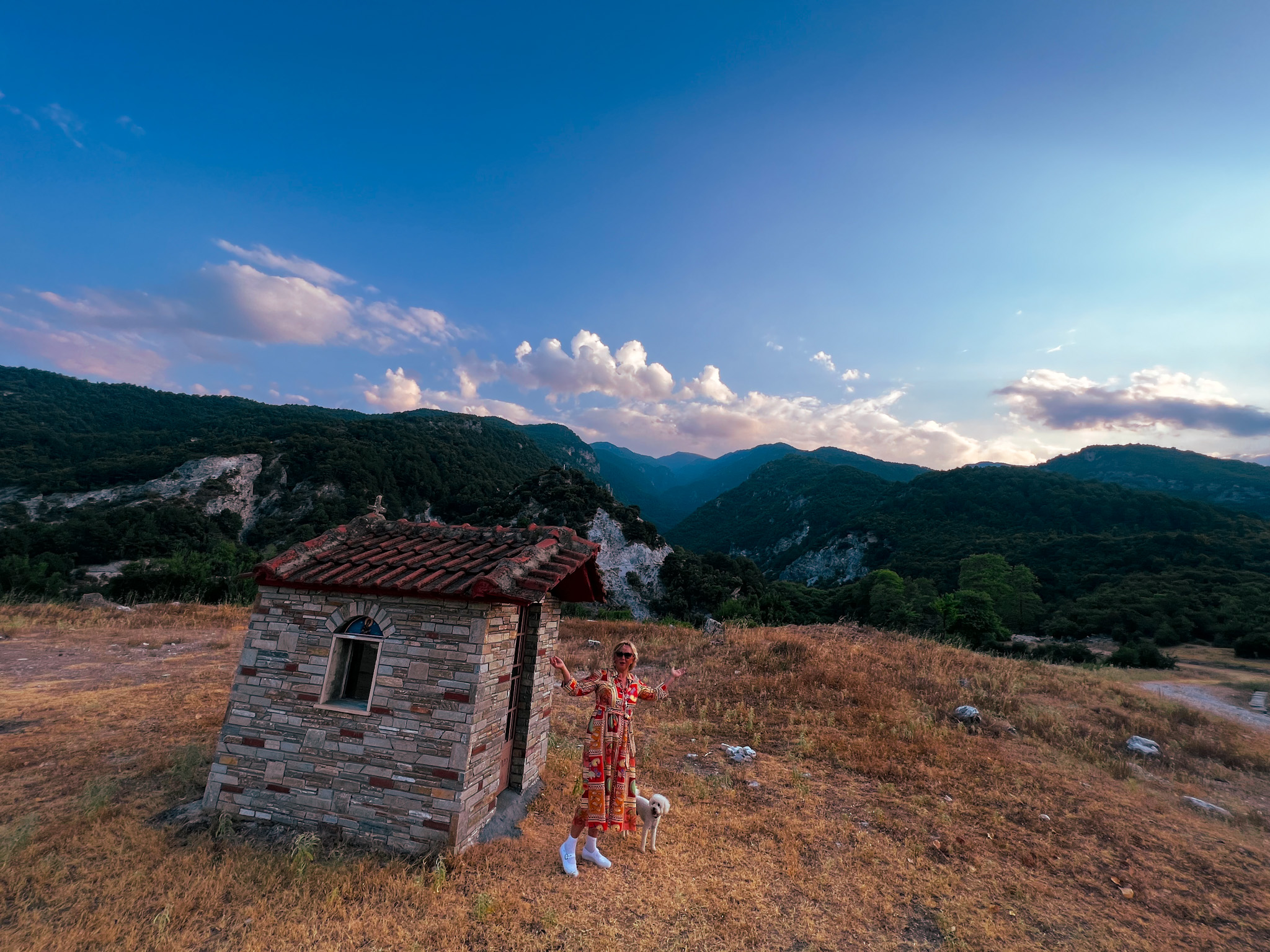 A person wearing a colorful, patterned dress stands with arms outstretched beside a small stone building with a red-tiled roof, in a dry mountainous area with lush green hills in the background. A small dog is standing nearby on the grass. The sky is clear with scattered clouds.