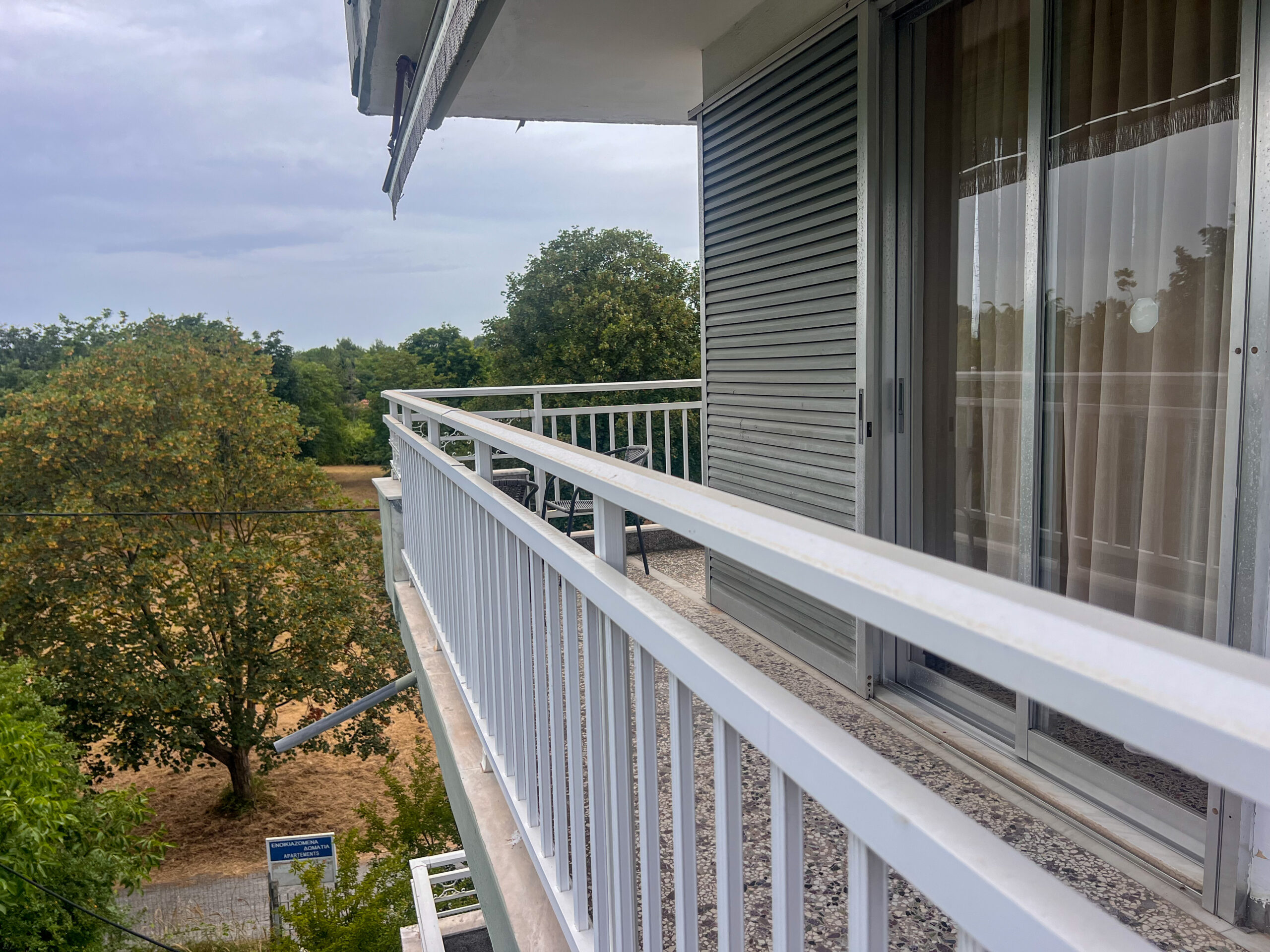 White-railed balcony connected to a room with sliding glass doors, overlooking a garden with trees