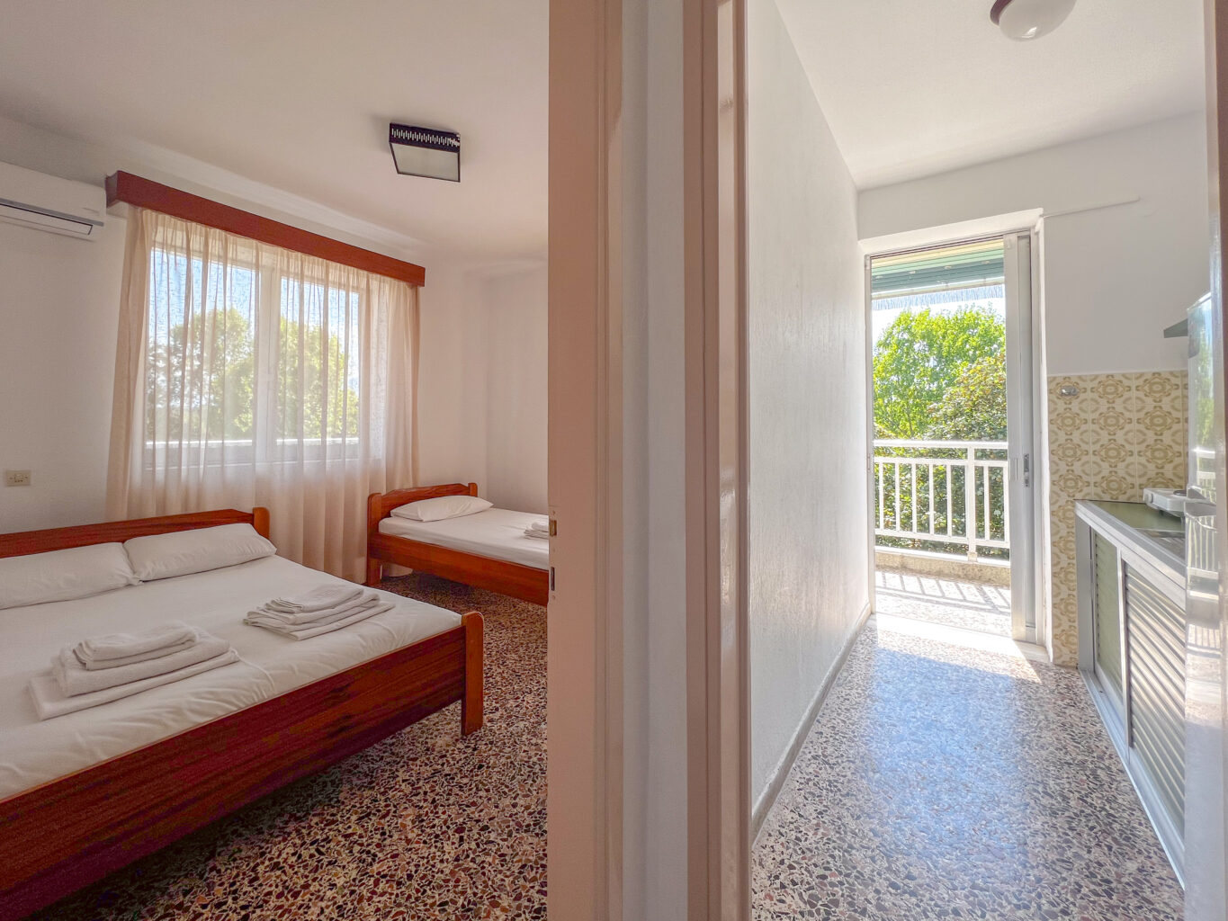 Hotel room with double and single beds, connected to a small kitchen with balcony access and views of the surrounding greenery.