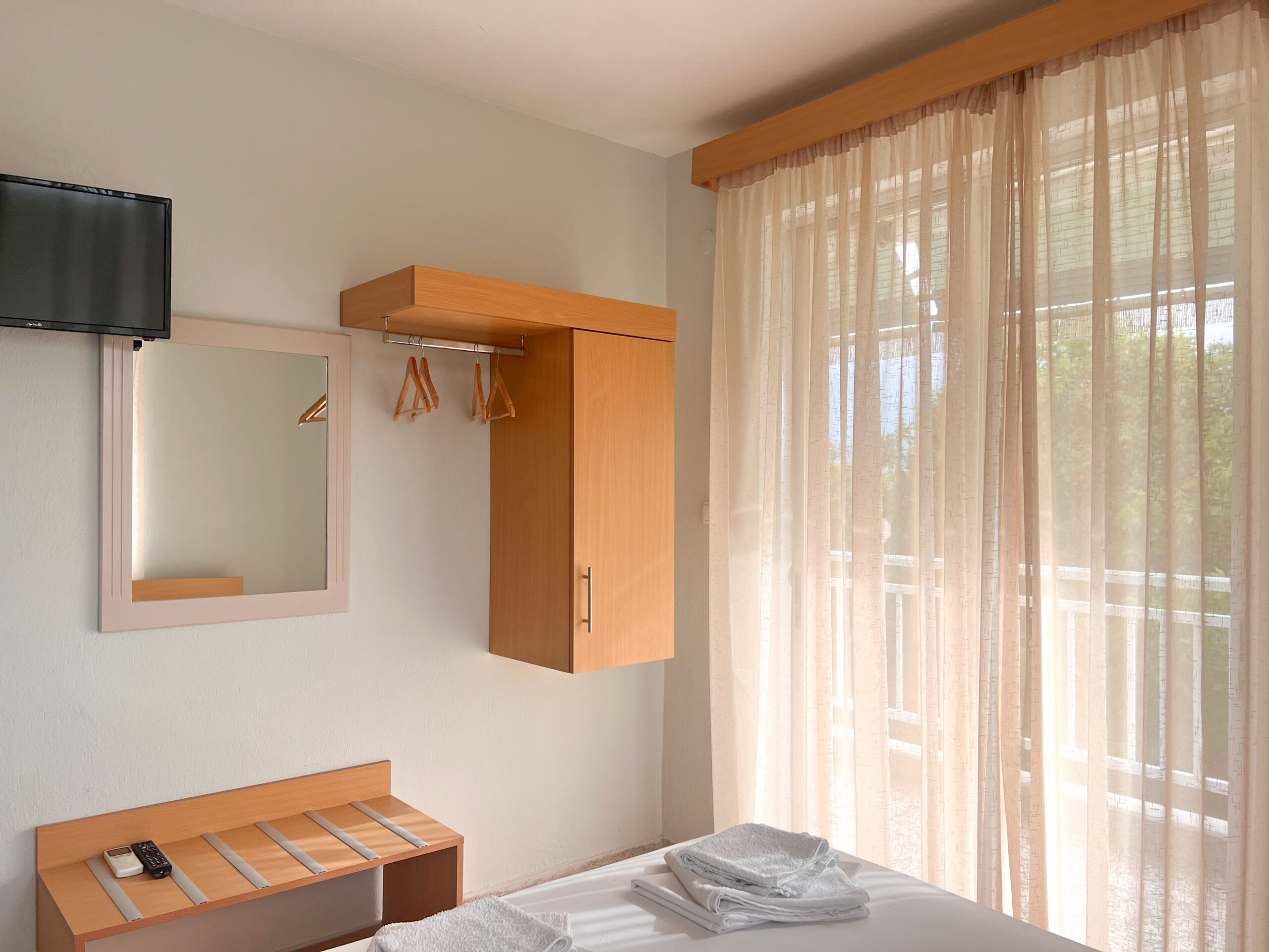 Hotel room with a TV, mirror, wooden wardrobe, and sunlight coming through large curtains