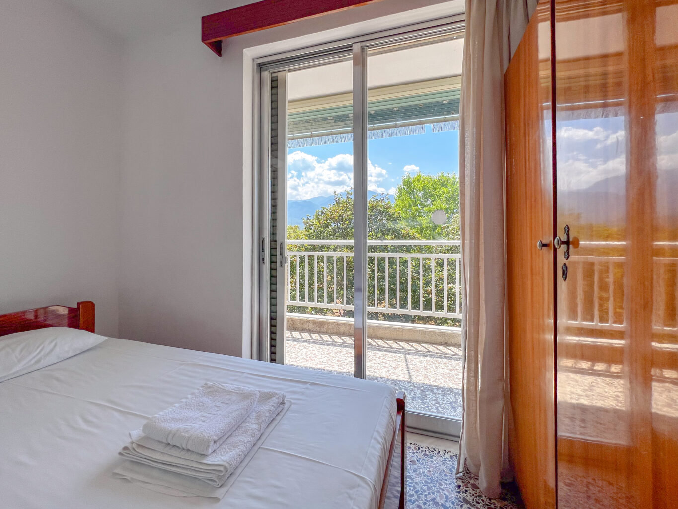 Hotel room with a wooden wardrobe, small dresser, TV, and large windows leading to a balcony with garden views.