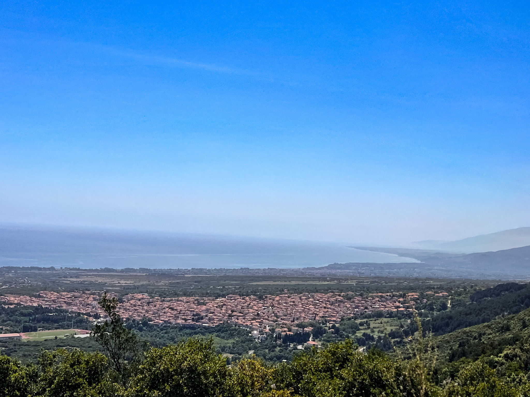 A panoramic view of the town of Litochoro nestled among green hills, with the coastline and the sea stretching into the distance under a bright blue sky.