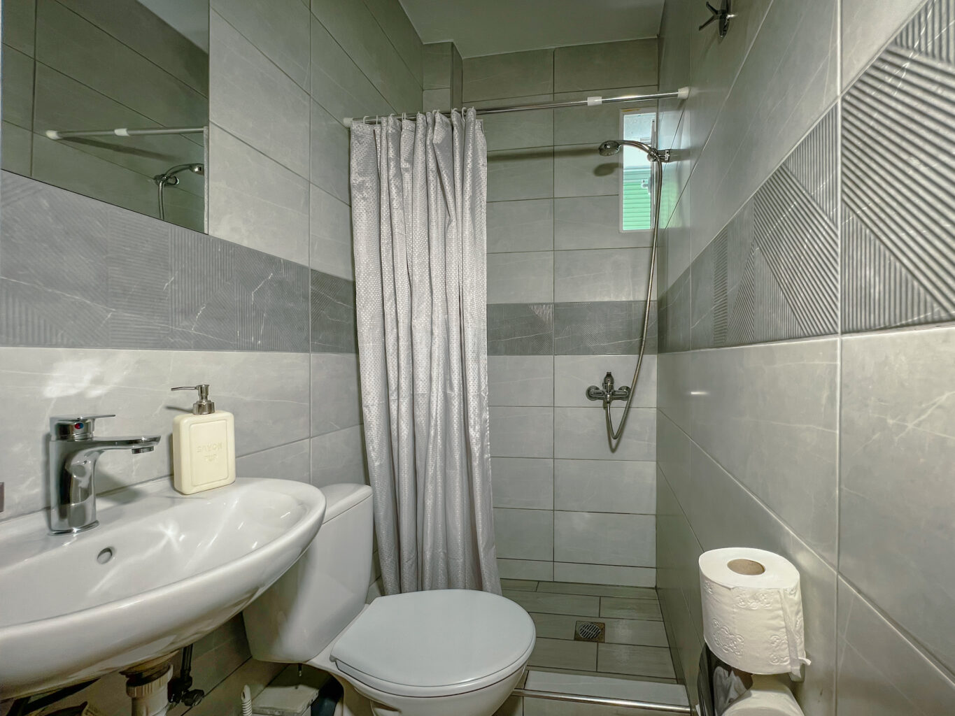 A small bathroom featuring a white sink, toilet, and a shower area with a gray and white tiled design. The shower is separated by a curtain and includes a handheld showerhead, with geometric tile patterns on the wall.