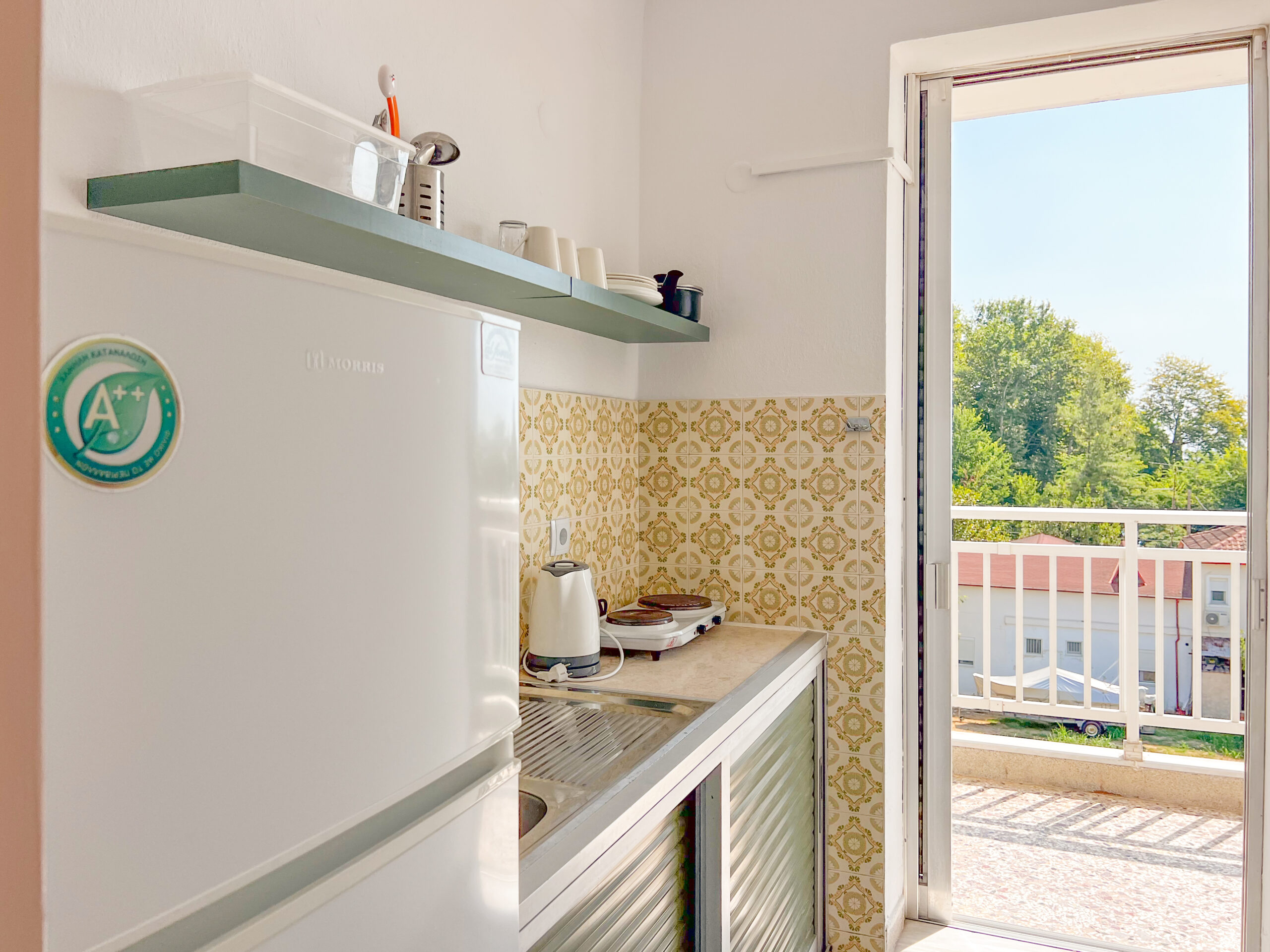 Small hotel kitchen with a refrigerator, shelves, and access to a balcony with a view of the surrounding greenery