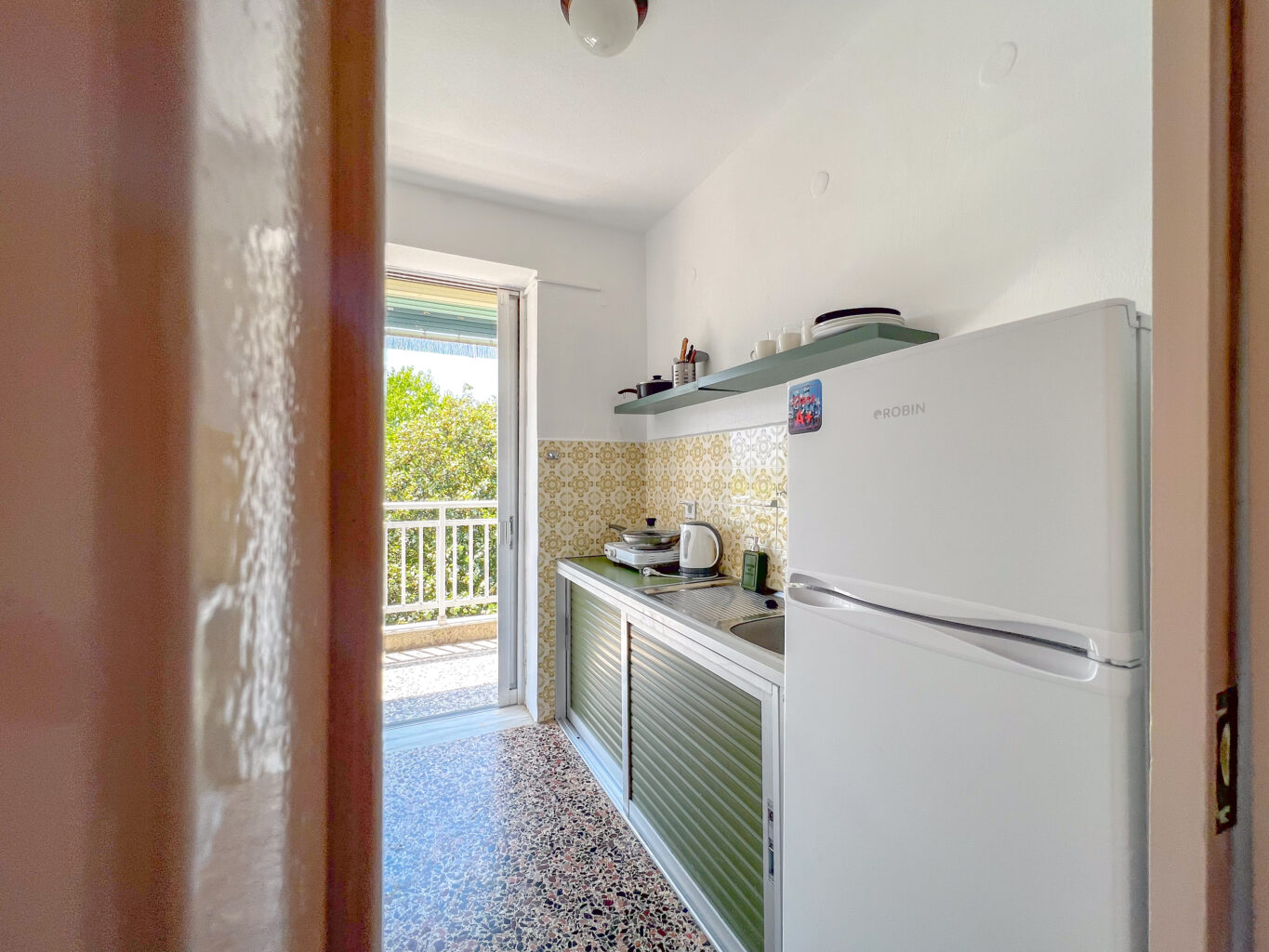 Small kitchen with a refrigerator and stove, leading to a balcony with a view of trees and greenery.