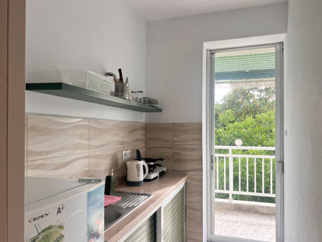 A modern kitchenette in Prado Hotel Apartments featuring a small refrigerator, microwave, electric kettle, and a neatly arranged shelf with kitchen utensils, overlooking a balcony with a lush garden view