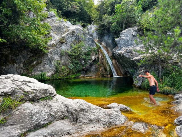 A man steps carefully into a stunning natural pool with a small waterfall, surrounded by rugged rocks and lush greenery, captured in the wilderness near Prado Apartment Hotel.