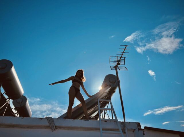 A woman in swimwear poses on the rooftop next to solar water heaters under a clear blue sky, demonstrating Prado Hotel Apartments' commitment to sustainable and eco-friendly practices.