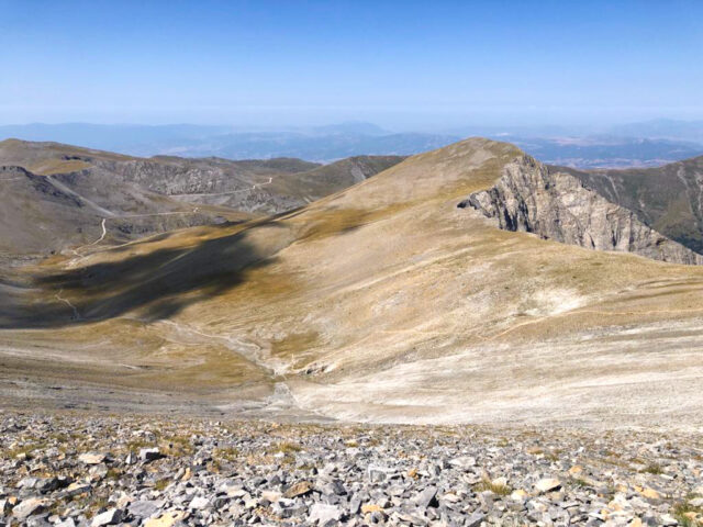 A panoramic view of the barren yet striking landscape of Mount Olympus, featuring rocky terrain and sparse vegetation under a clear blue sky, with distant mountains fading into the horizon.