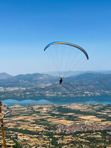 A paraglider soars above a vast landscape showcasing the lush fields, winding roads, and the serpentine lakes near Mount Olympus under a clear blue sky.