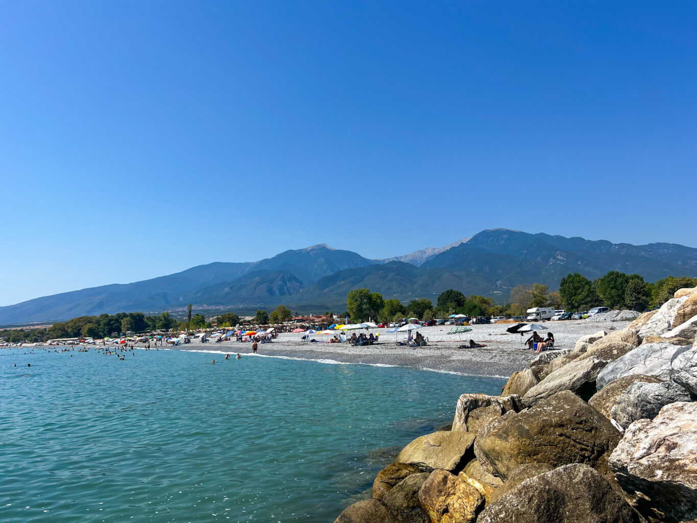 View of Gritsa Beach with people relaxing on the shore, Mount Olympus in the background, and clear blue water.