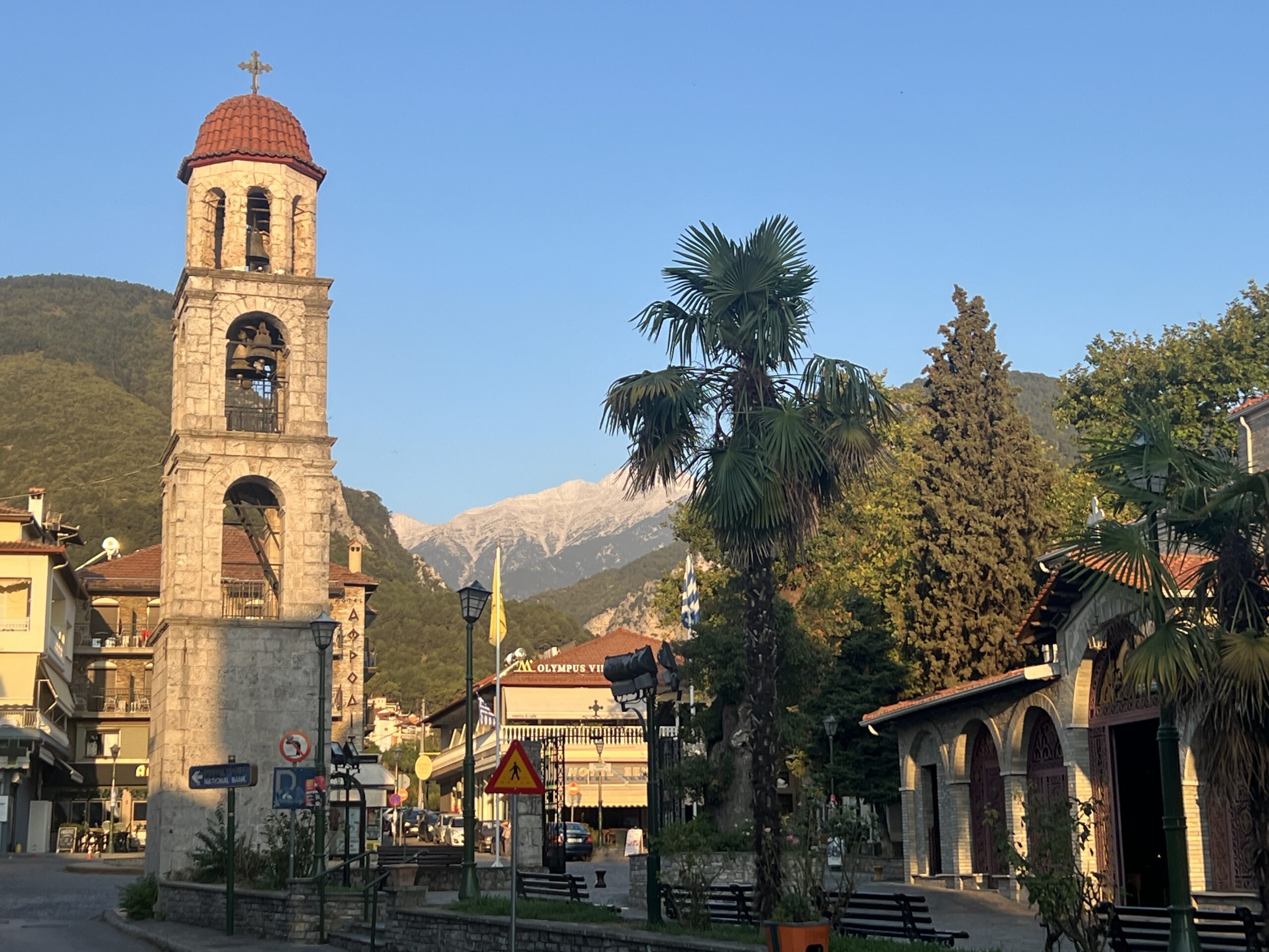 A view of Litochoro with its iconic stone bell tower and a background of Mount Olympus, surrounded by lush greenery and palm trees under a clear blue sky.