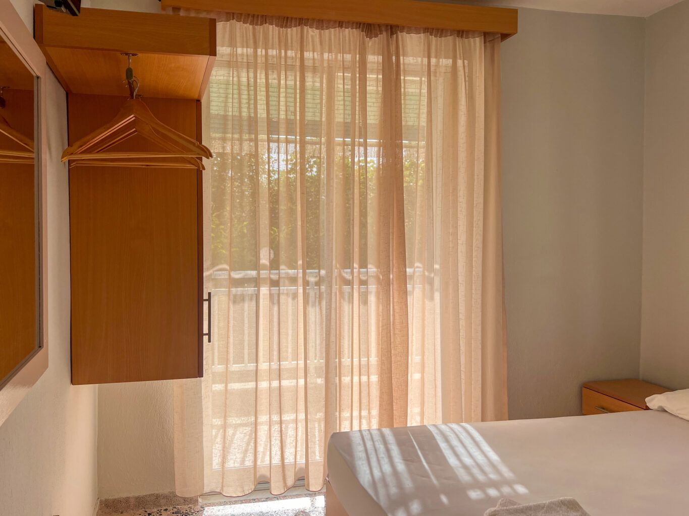 Bright hotel room with a wooden closet, hangers, and large curtains letting in sunlight from the balcony.