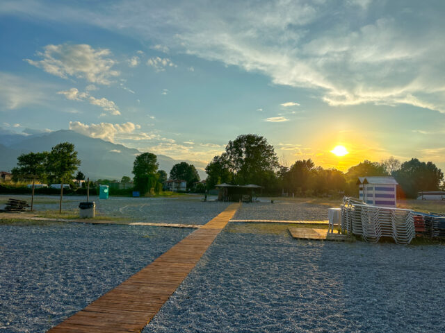 A wooden boardwalk leading to a tranquil beach at sunset, with the sun casting golden hues over the pebbly shore and Mount Olympus in the background, captured at Gritsa Beach.