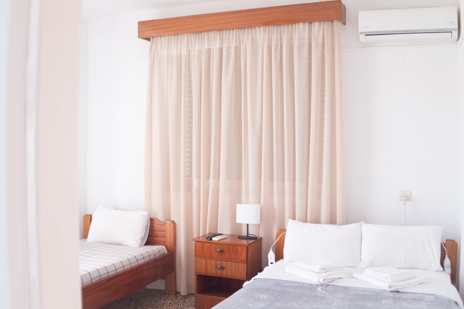 Interior view of Room 32 at Prado Apartment Hotel with a double bed and single bed, soft curtains, towels, and natural light.